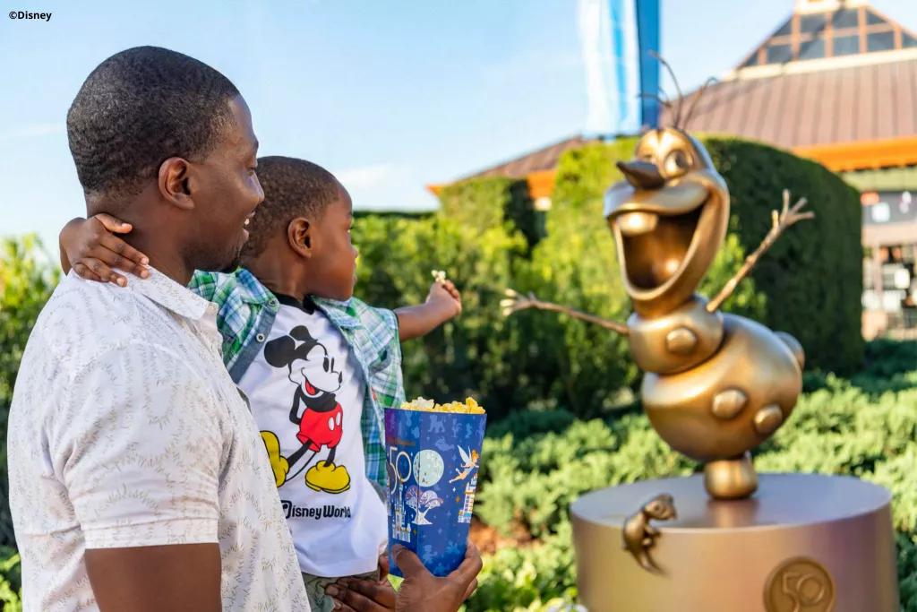 A man holding his son pointing towards the golden Olaf statue in EPCOT