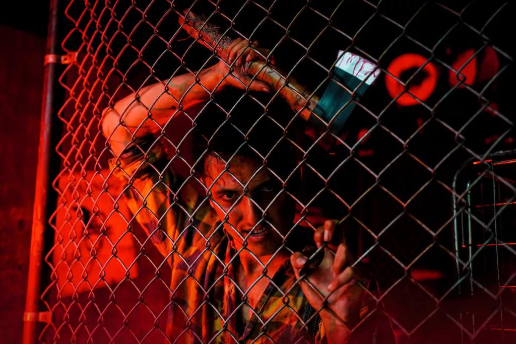 A man with an axe leaning up against a chain fence in a dark room lit with red lights