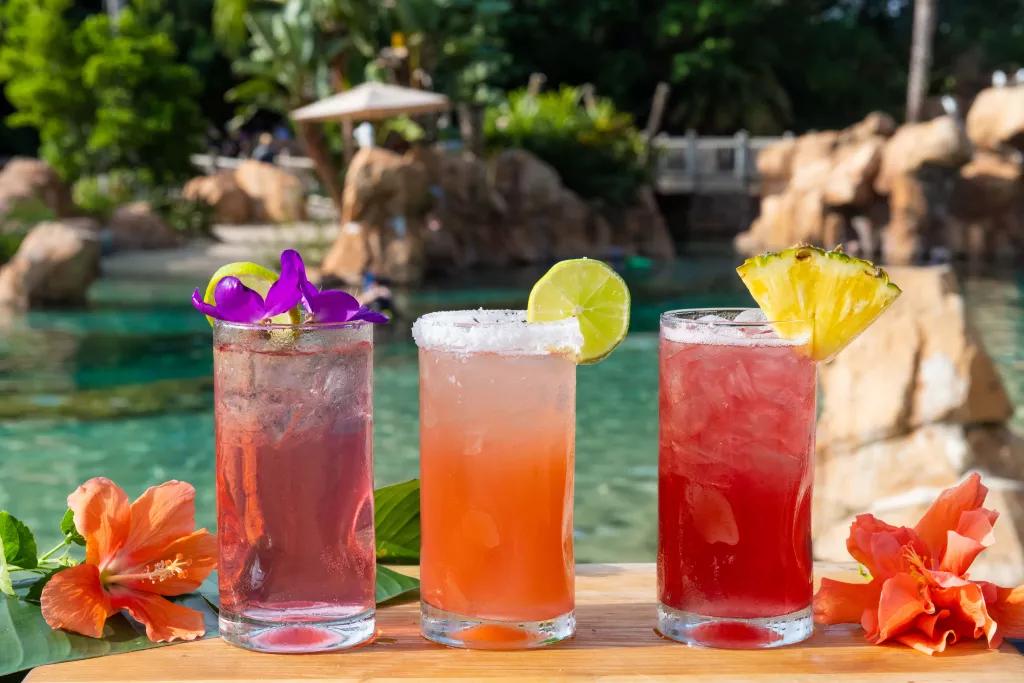 Three fruity cocktails surrounded by flowers in front of a beach