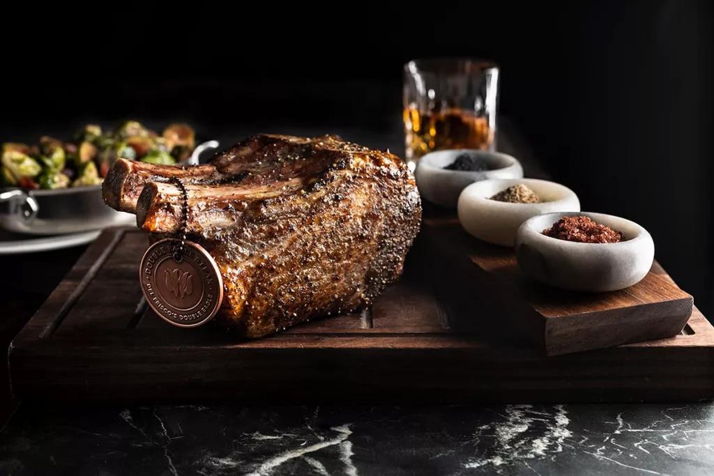 A cut of cooked steak on a wooden board with pots of spices and a glass of whiskey