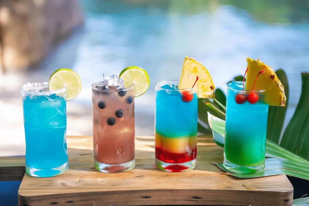 Four colourful cocktails on a wooden board in front of a sandy beach