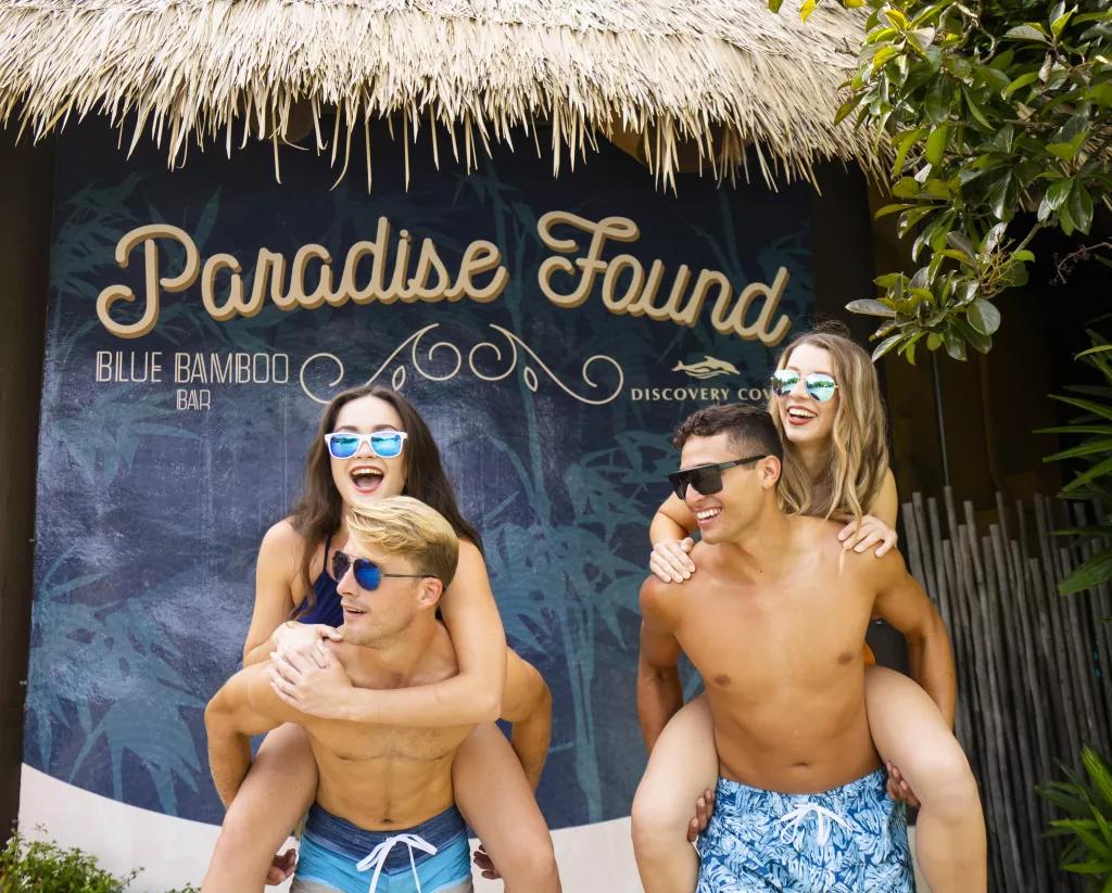 Two men in swimming trunks giving two women piggy backs in front of a wall that says "Paradise Found"