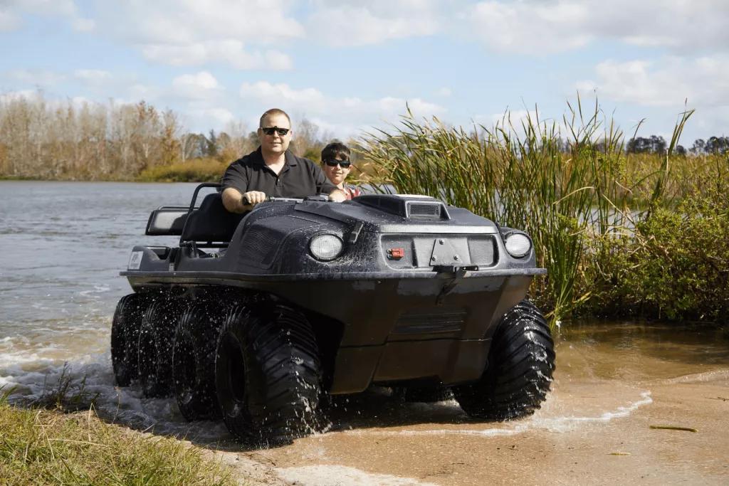 A man and boy sitting in a Mucky Duck off road vehicle in front of a lake