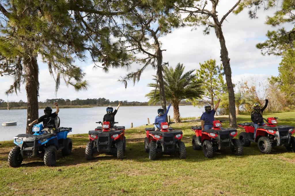 5 people riding 4x4 ATVs posing for the camera in front of a lake with one fist in the air