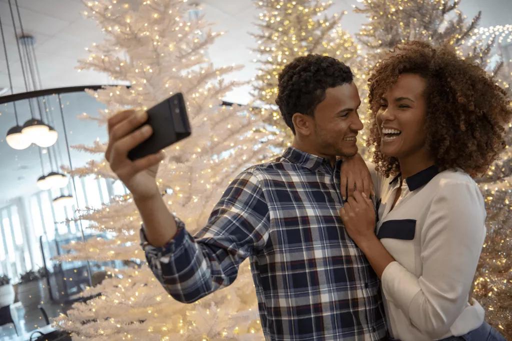A man and a woman smiling while taking a selfie in front of a lit up Christmas tree