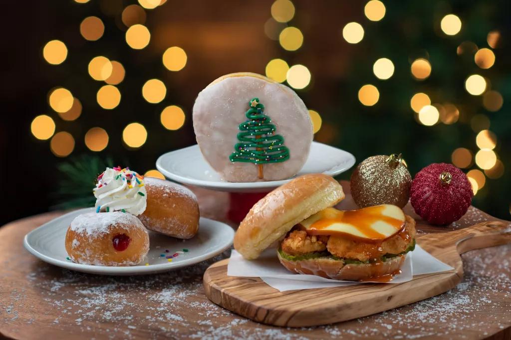 Plates of food with a chicken sandwich and doughnuts in front of Christmas trees and lights