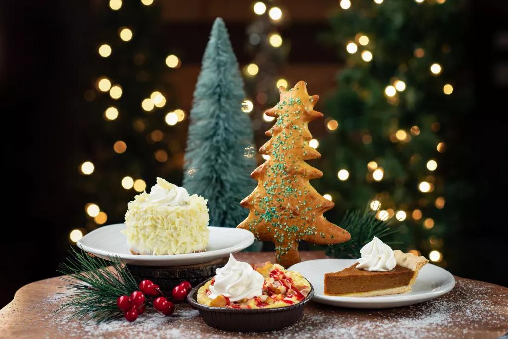 Plates of food including pumpkin pie and an iced cake infront of Christmas trees and lights