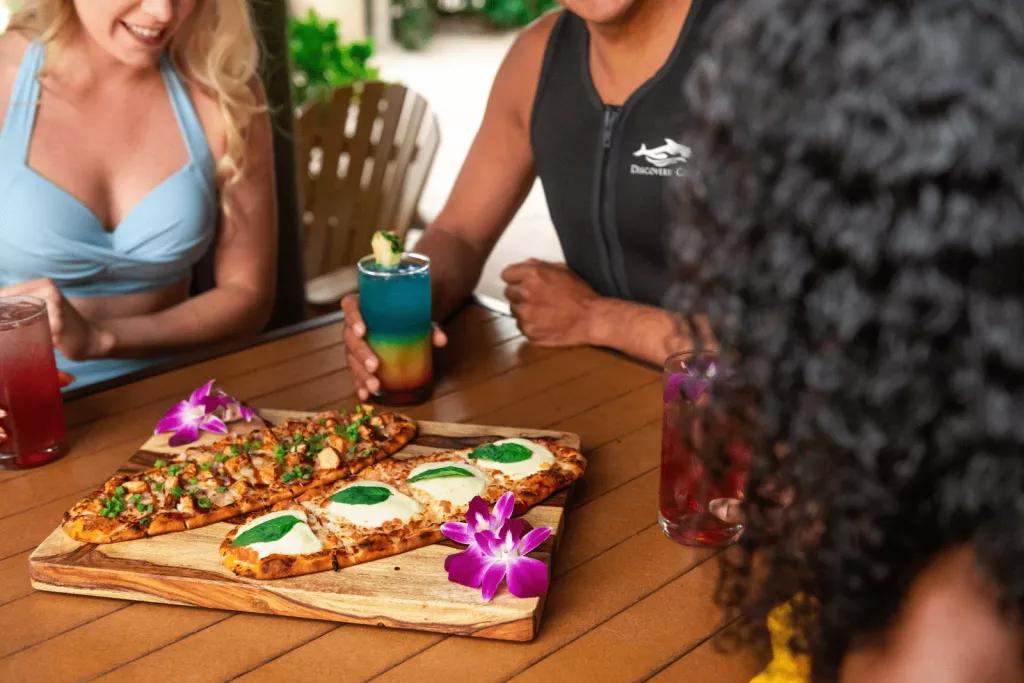 Three people in wetsuits and swimming costumes sitting around a wooden table with two pizzas on it