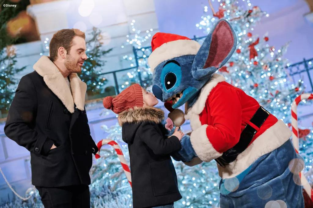 Stitch dressed as Santa being kissed on the nose by a small child. Christmas trees and candy canes are in the background