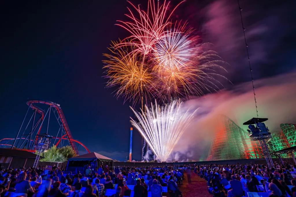 A large crowd of people watching fireworks go off above a rollercoaster