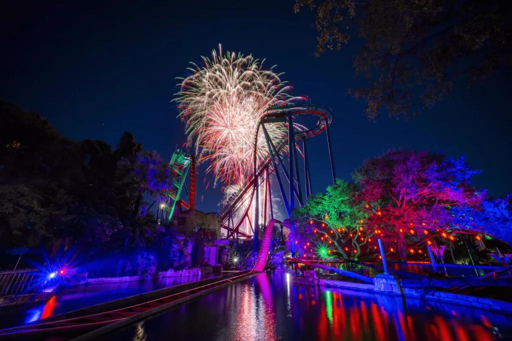 Large colourful fireworks in a dark sky, behind a tall rollercoaster and some trees 