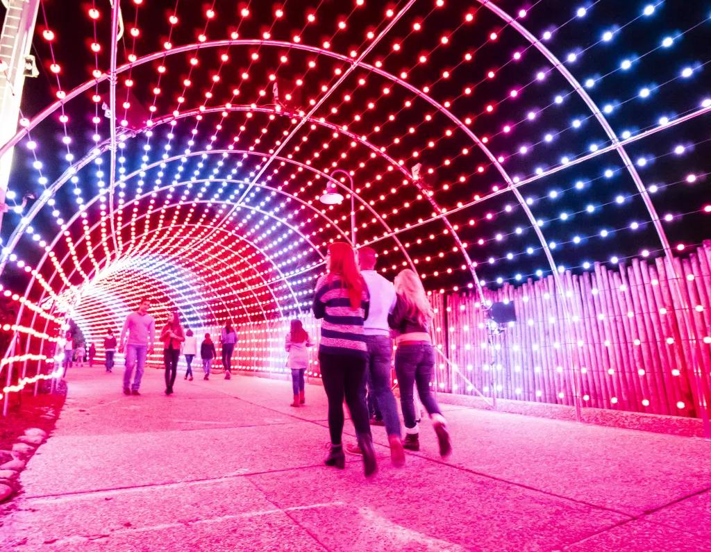 A crowd of people walking away from the camera through a tunnel lit by multi-coloured LEDs