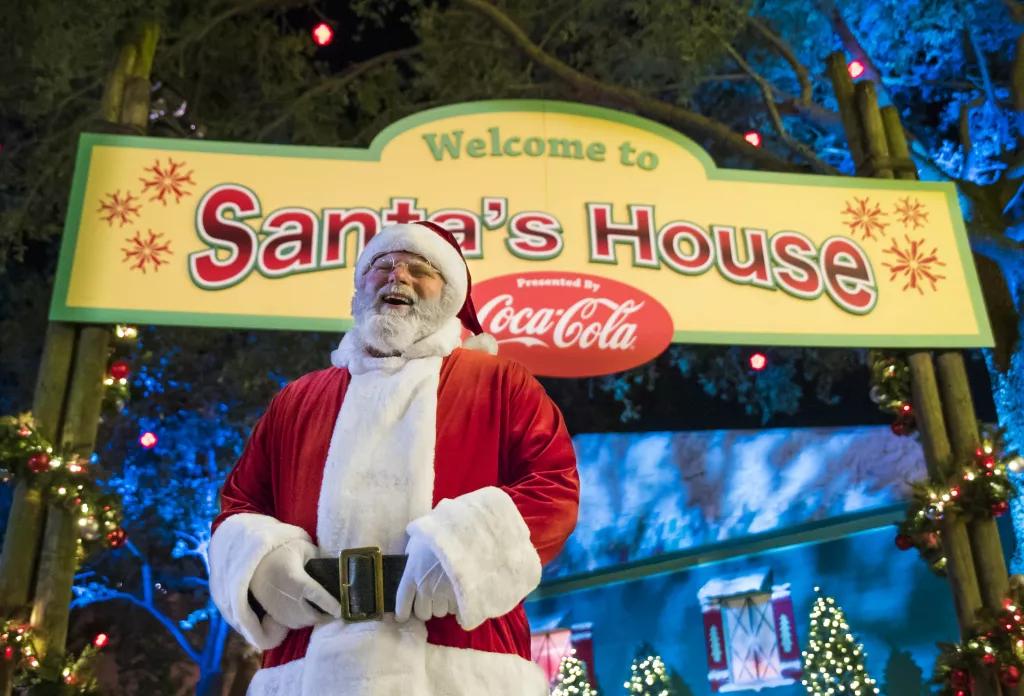 Santa Clause laughing while standing in front of a sign that says "Welcome to Santa's House"