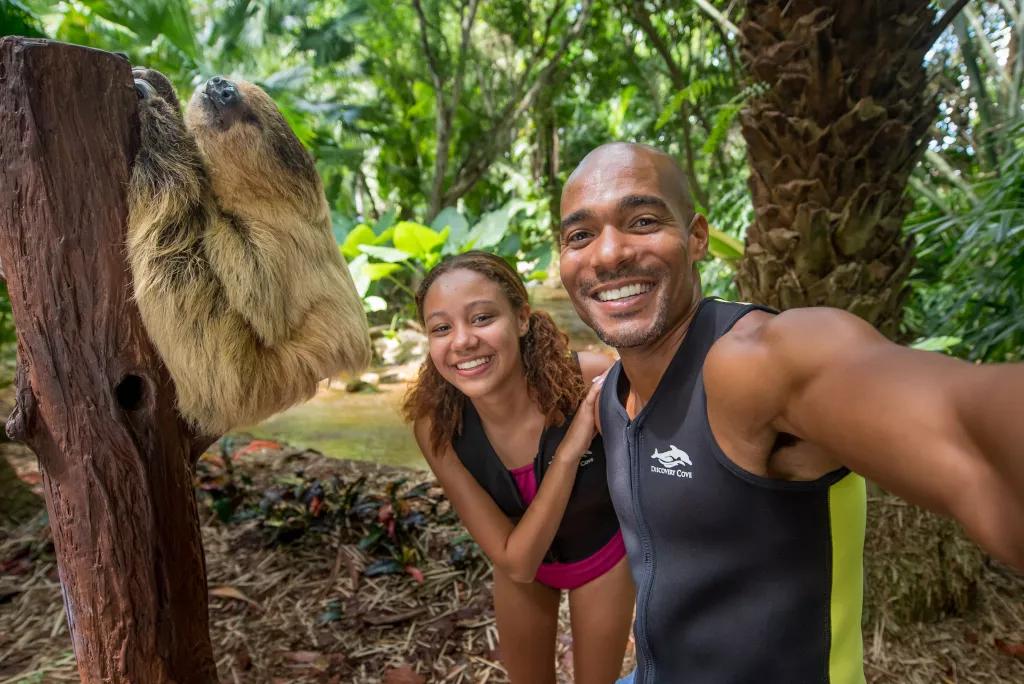 A man and a woman taking a selfie with a sloth climbing a tree stump
