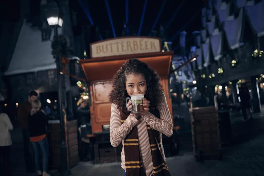 A girl wearing a red a yellow scarf drinking a hot butterbeer in front of a Butterbeer cart