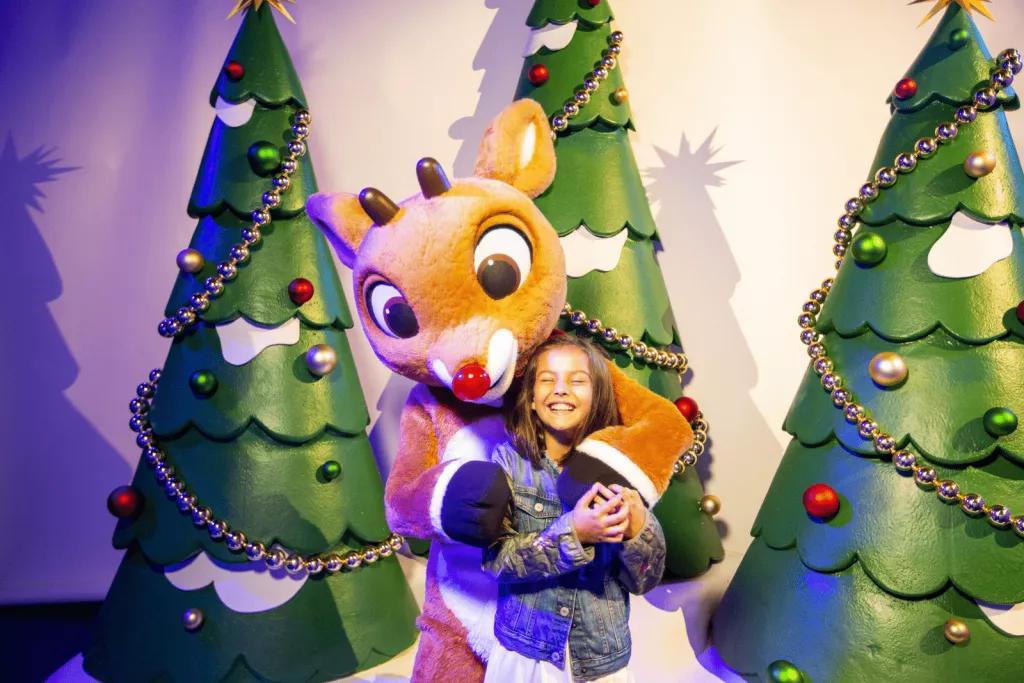 A child being hugged by someone in a Rudolph costume, with cartoon Christmas trees behind them