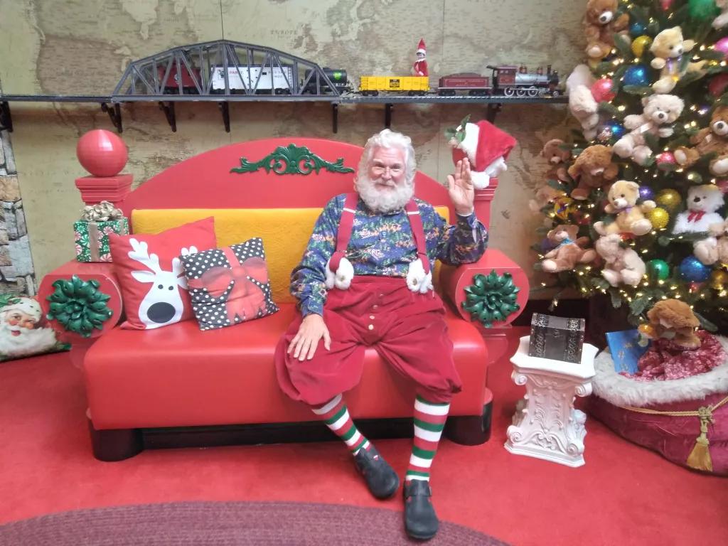 Santa Claus waving at the camera while sat on a sofa next to a Christmas tree and Christmas presents