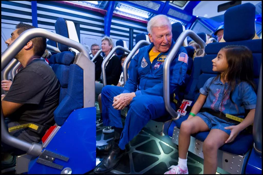 An astronaut wearing a blue jumpsuit sat next to a young girl, both strapped into a simulator