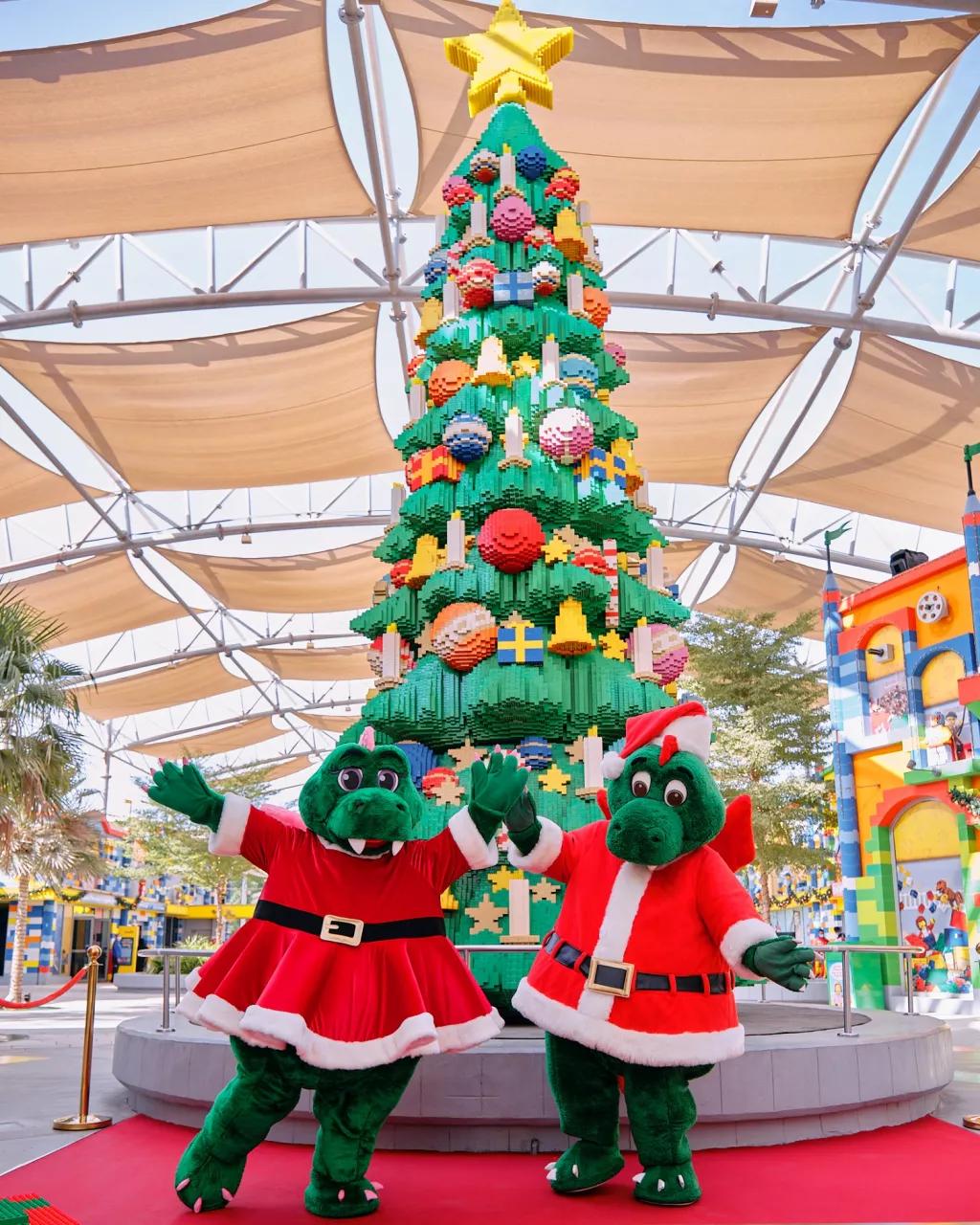 Two crocodile characters dressed as Santa and Mrs Claus standing in front of a LEGO Christmas tree