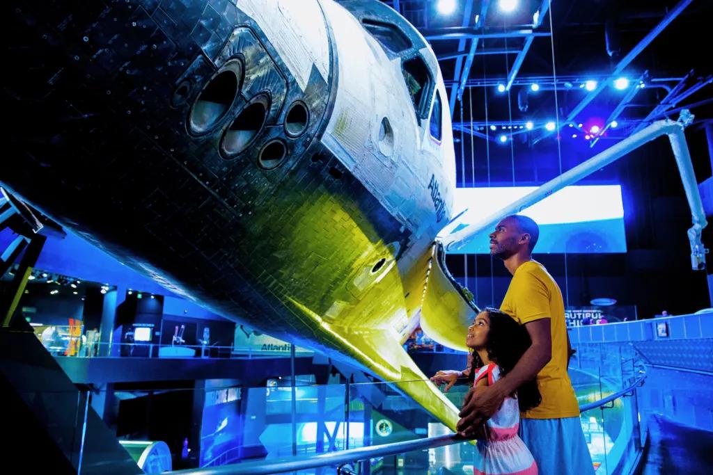 A father with his arm around his young daughter looking up at the Space Shuttle Atlantis