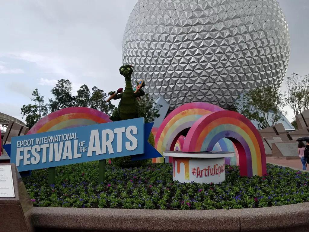 A topiary of Figment the dragon in front of Spaceship Earth with a Festival of the Arts sign