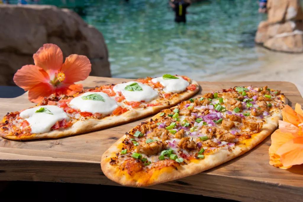 Two long flatbread pizzas on a wooden board in front of a the clear blue waters of the beach