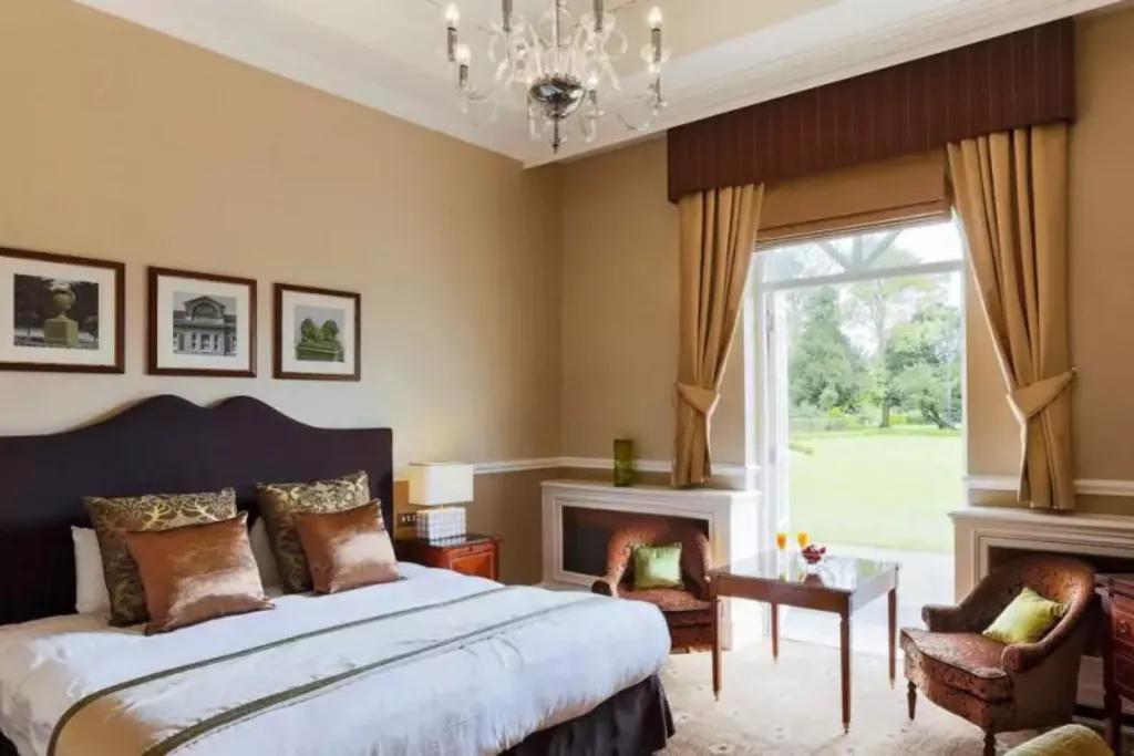 A hotel room with a bed and two armchairs with open French doors looking out to a grassy garden