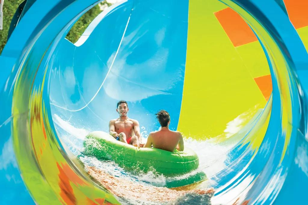 Two men in swimming costumes riding in a rubber raft down a multicoloured waterslide