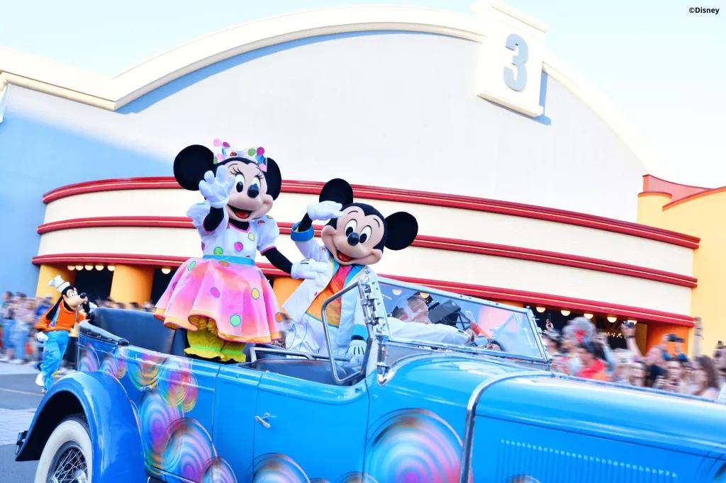 Mickey and Minnie Mouse wearing rainbow outfits waving at a crowd from a convertible car