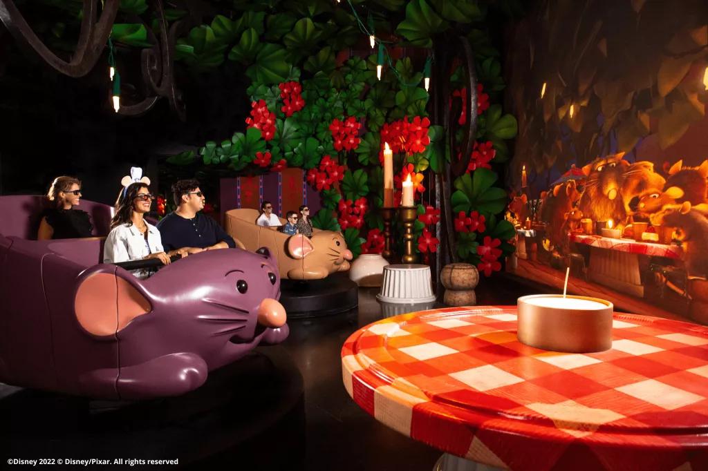Guests in ride vehicles shaped like rats looking at a large screen and wearing 3D glasses. They are surrounded by larger than life objects