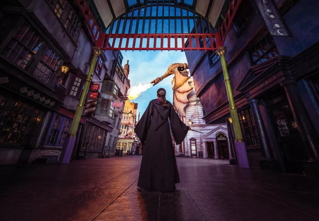 A girl in Diagon Alley wearing wizard robes and holding a wand, looking up to a fire breathing dragon on the top of a building