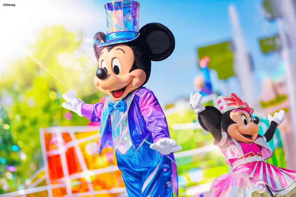 Mickey and Minnie Mouse in bright coloured iridescent clothes in front of a parade float