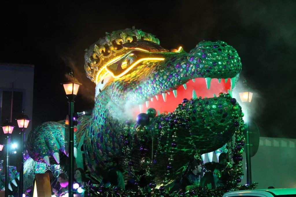 A large parade float shaped like an alligator wearing a crown and holding strings of beads in its mouth