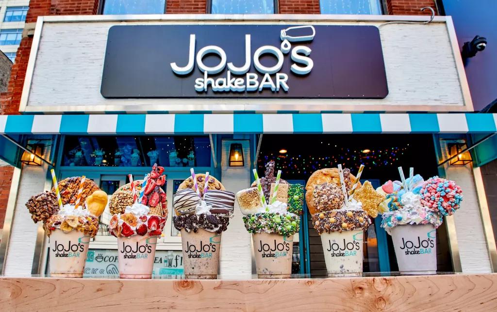 A line of large milkshakes with whipped cream, sprinkles and cookies on top in front of a JoJo's Shake Bar sign
