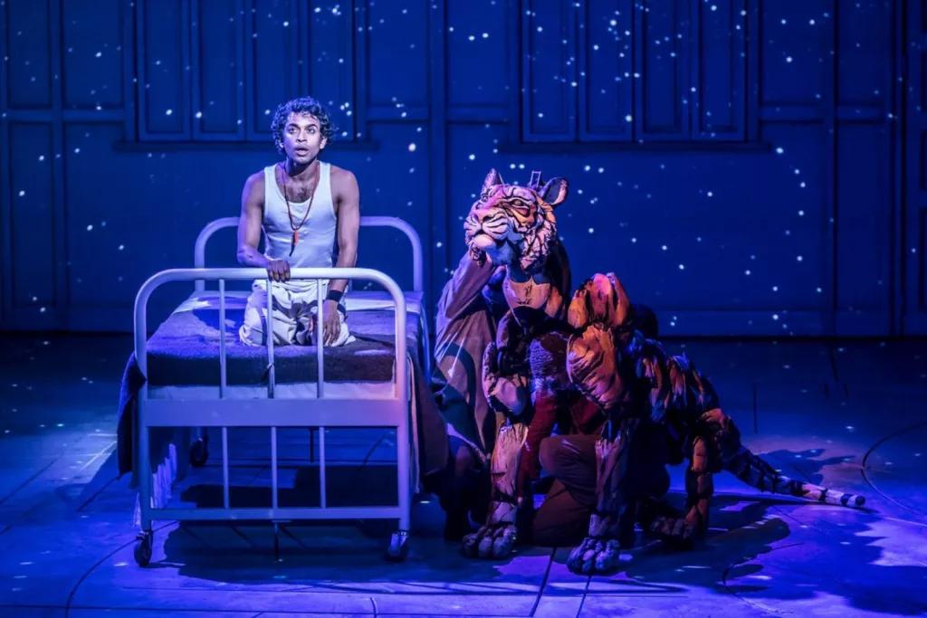 A man on a stage kneeling on a single metal-framed bed next to a life-sized puppet of a tiger