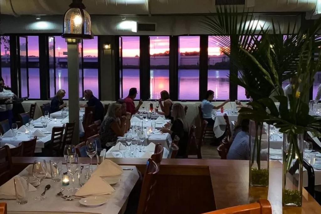A restaurant dining room with people eating at tables covered with white table cloths. There is a sunset outside reflected on the water, turning it a light purple.