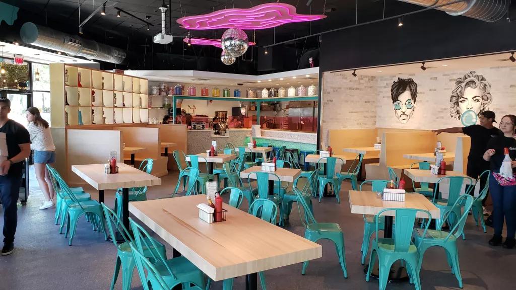 The interior of a diner style restaurant with light wooden tables and teal chairs