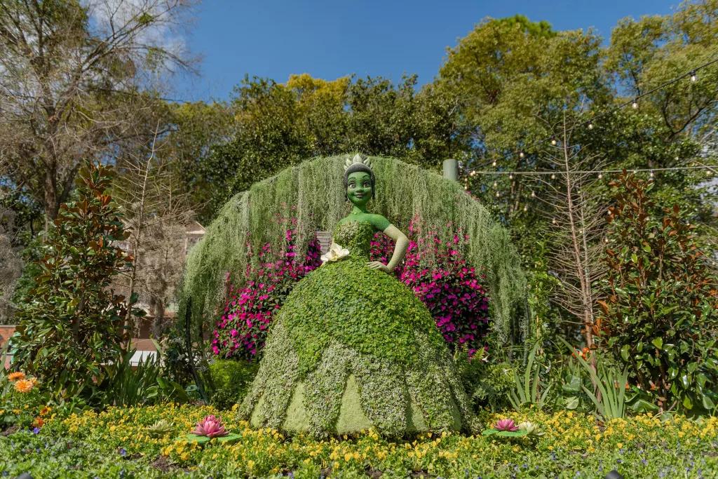 A life-size topiary designed to look like Princess Tiana in front of a wall of plants and flowers