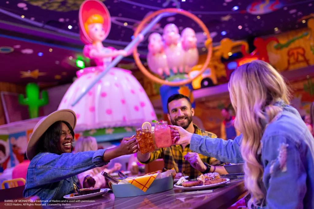 3 people sat at a table clinking mason jars full of drinks. They are sat in front of a large figure of Bo Peep from Toy Story