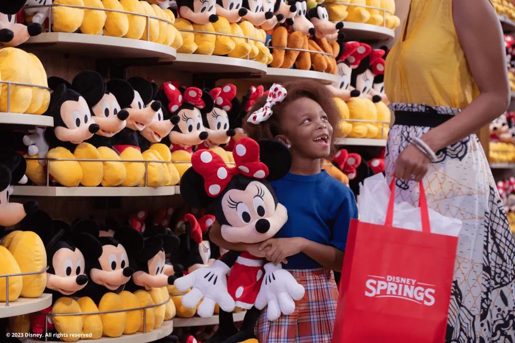 A young girl holding a large Minnie Mouse plush looking up at a woman. They are standing in front of a wall of Disney plushes