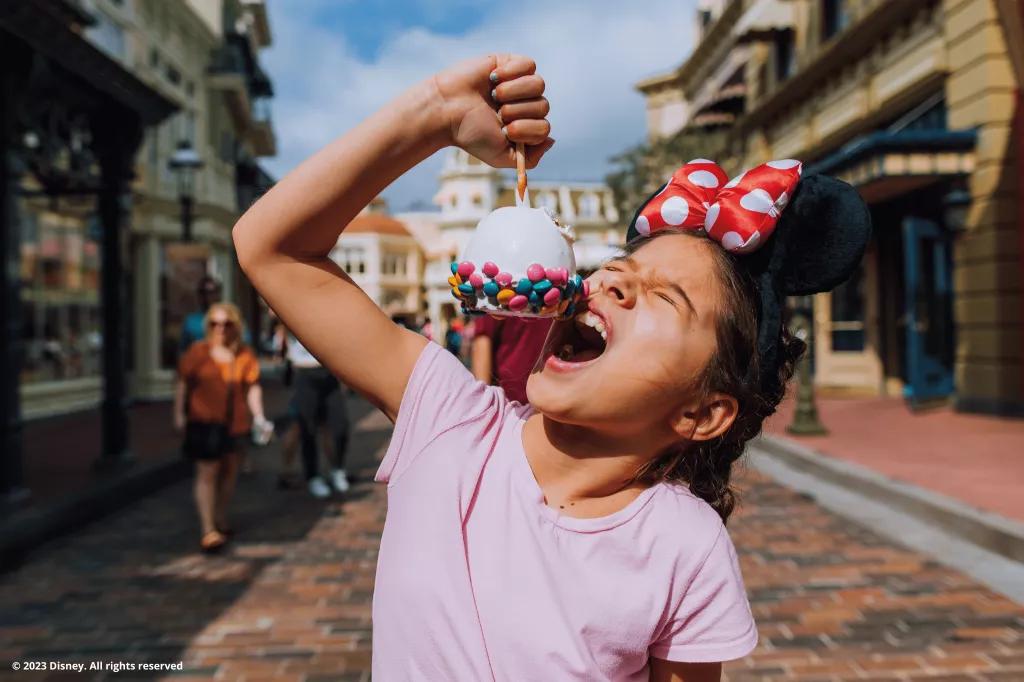 A young girl wearing a pink tshirt and Minnie Mouse ears biting into a large candy apple