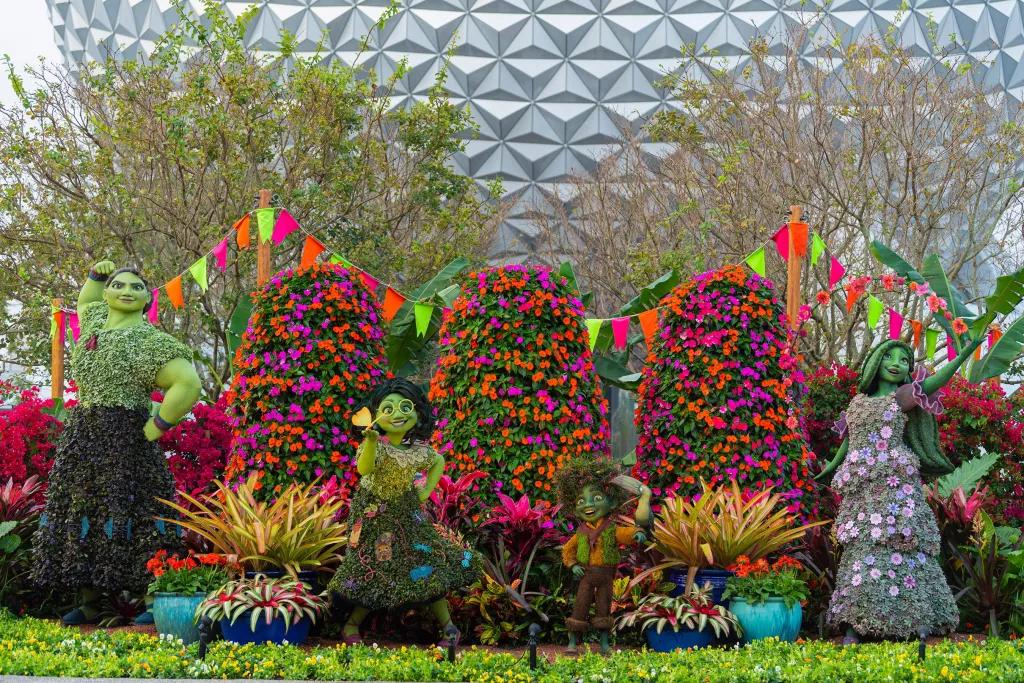 Topiaries shaped like characters from Encanto in front of colourful flowers and hedges with Spaceship Earth in the background