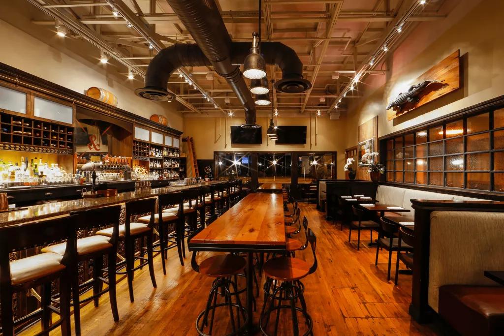 The interior of a restaurant with wooden table and floors and a long bar with stools along one side