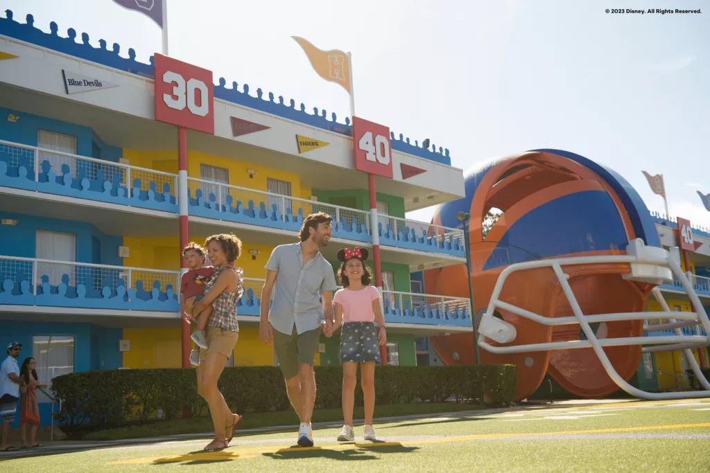 A family of four walking past a motel-style hotel building on grass designed to look like an American football field. There is a giant American football helmet in the background. 