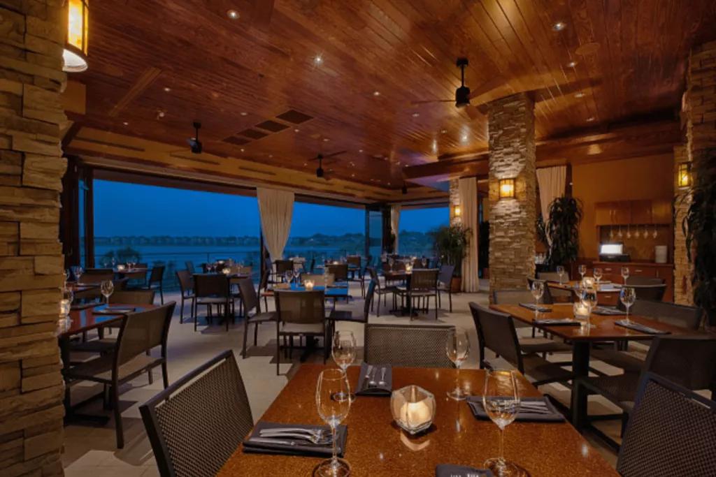 An empty restaurant filled with tables and chairs and a glass backwall looking out onto a lake