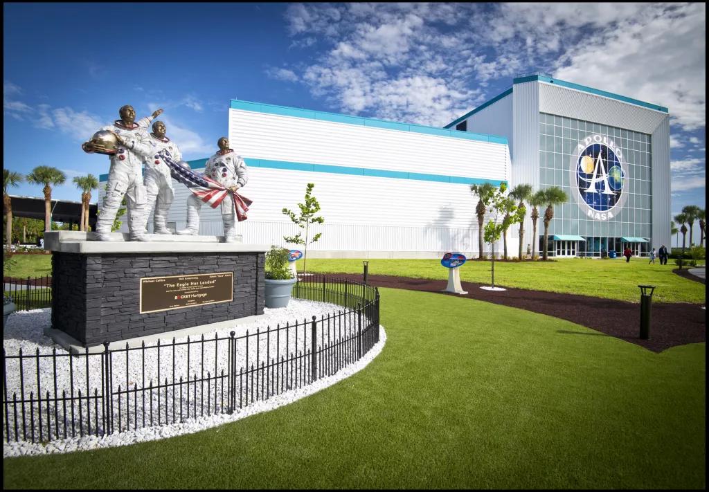 A statue of three astronauts holding an American flag in front of a building with a large sign reading Apollo NASA