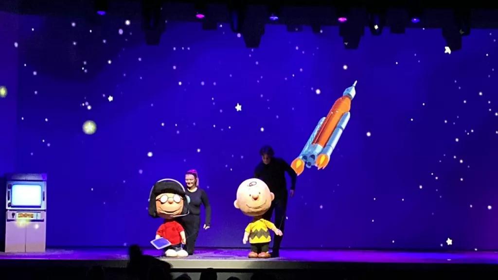 Two people on stage holding life-size puppets depicting Charlie Brown and Lucy from the Peanuts comics