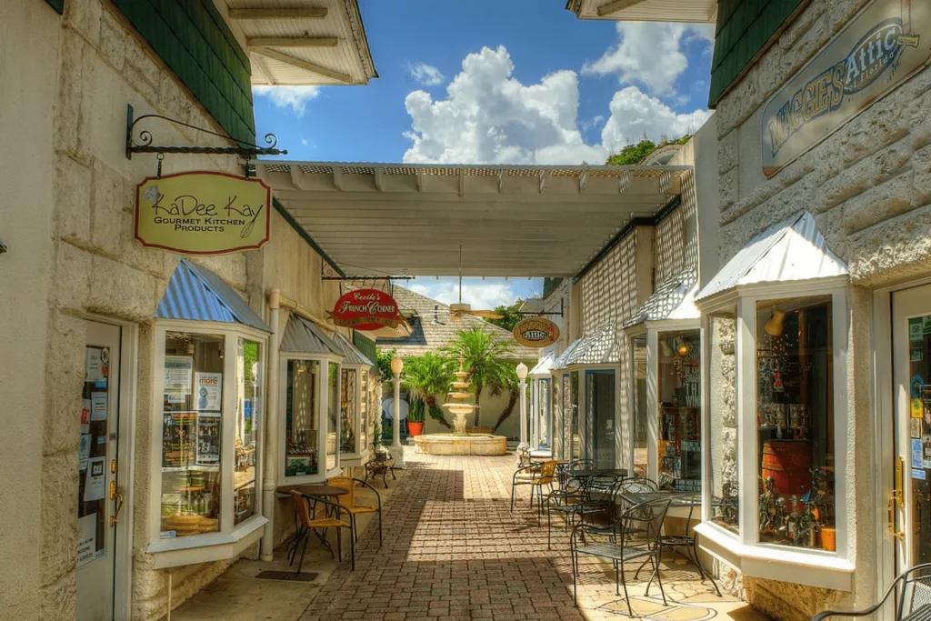 A covered walkway lined with shops with tables and chairs outside, leading to a fountain at the end