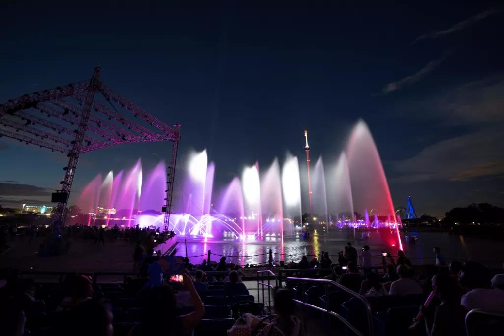 Fountains over a lake lit up at night, surrounded by spectators