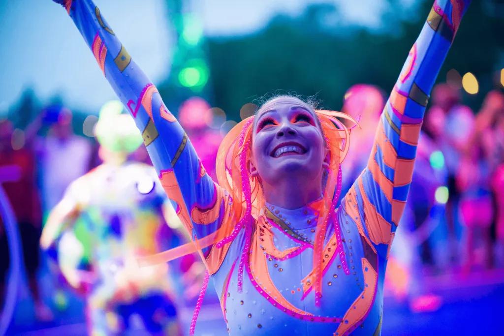A female performer in a neon blue and orange leotard looking up to the sky with her arms raised